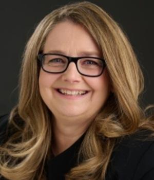 Smiling blond-haired woman wearing a dark top and dark-rimmed glasses